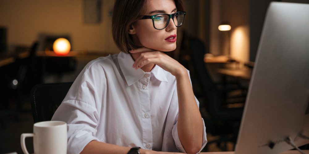 Serious young businesswoman in glasses sitting and working with computer in office